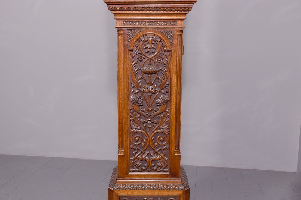 A late Victorian tall carved oak grandfather clock with a swan-neck pediment, arched brass dial, and extensive foliate relief carvings on the body and base. View X