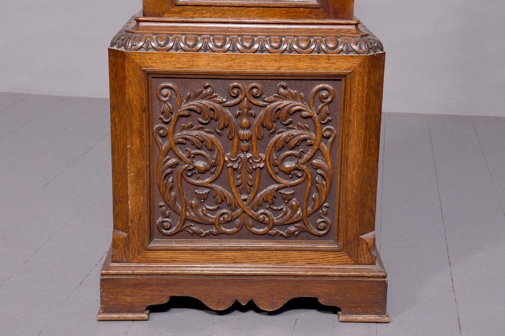 A late Victorian tall carved oak grandfather clock with a swan-neck pediment, arched brass dial, and extensive foliate relief carvings on the body and base. View XIII