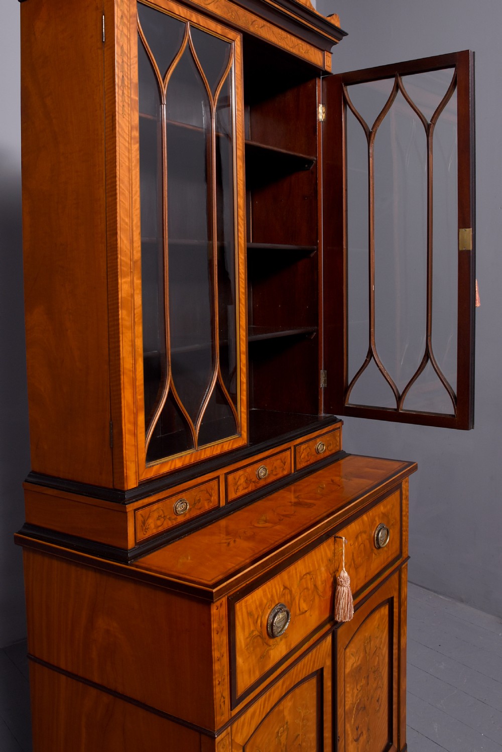 An antique satinwood secretaire bookcase featuring glazed upper doors with astragal tracery, detailed floral marquetry inlays, and an arched pediment. View XVI