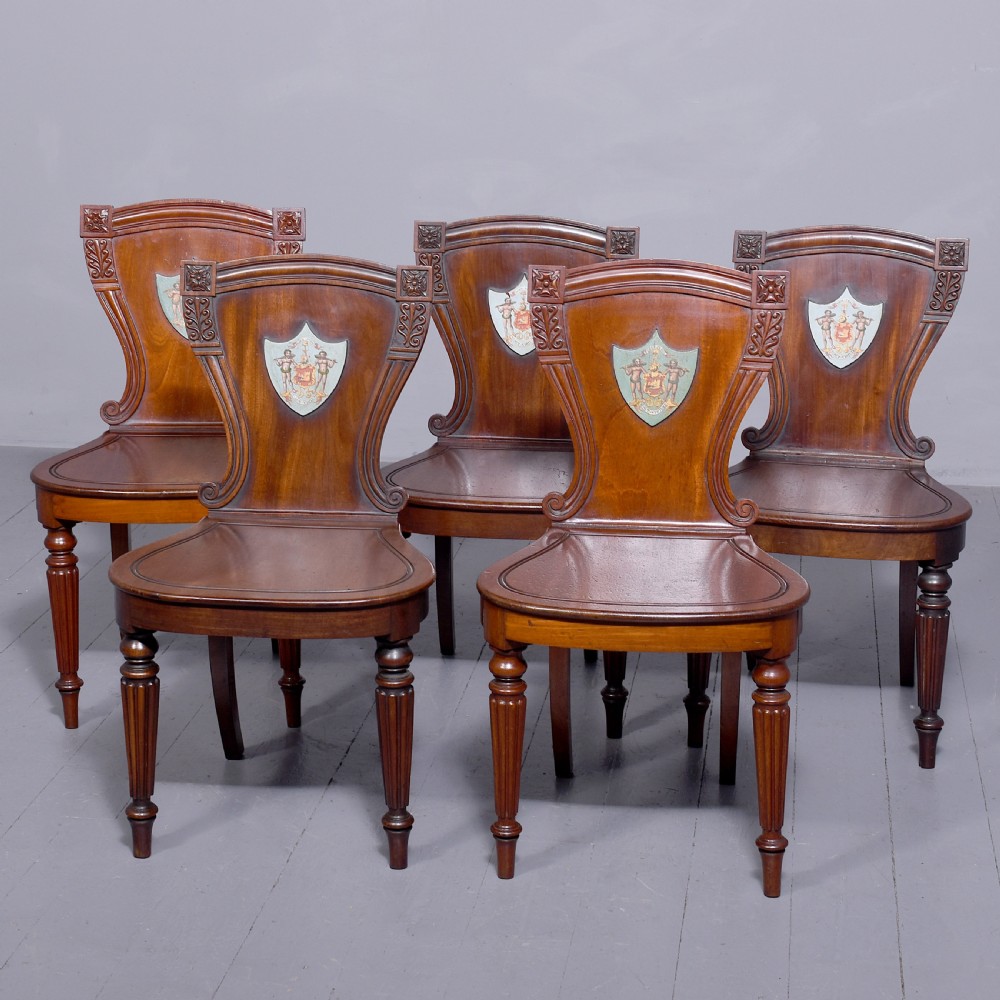 A set of five George IV mahogany hall chairs featuring carved backrest scrolls, turned fluted front legs, and hand-painted heraldic crest shields.