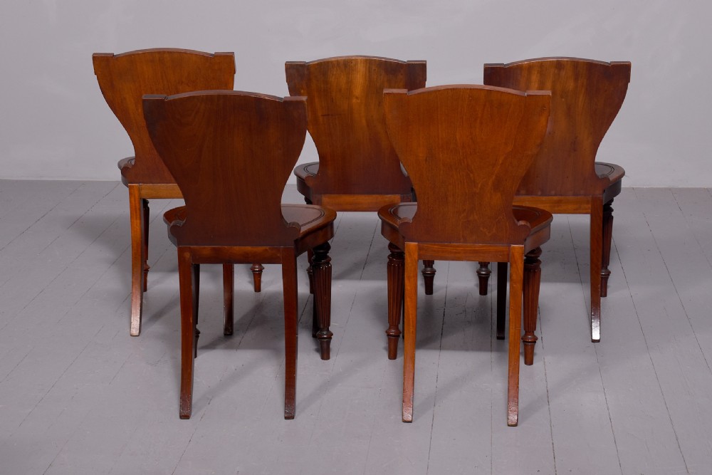 A set of five George IV mahogany hall chairs featuring carved backrest scrolls, turned fluted front legs, and hand-painted heraldic crest shields. View VII