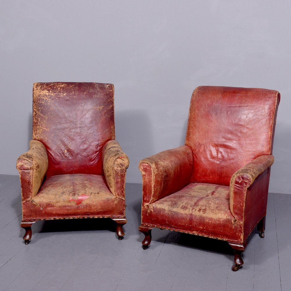 A pair of deep red leather library armchairs with high backs, rolled arms, brass stud trimming, and turned wooden front legs on castors.