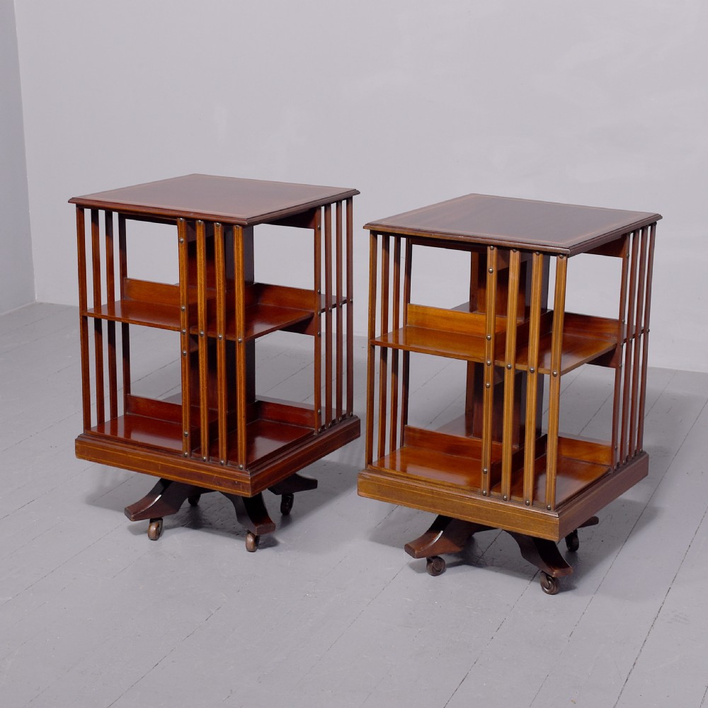 A pair of square Edwardian mahogany revolving bookcases featuring satinwood line inlay, open vertical slatted sides, two shelf tiers, and quadform bases on brass castors.
