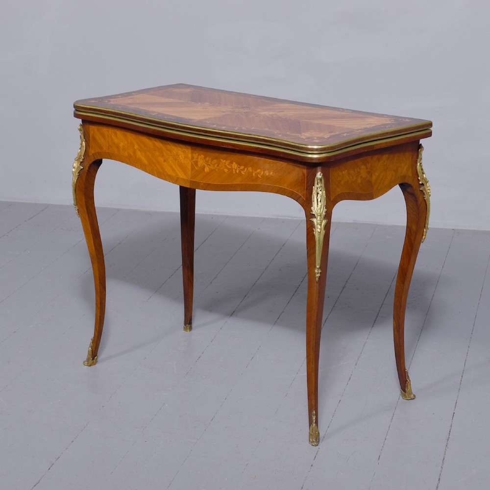 A Louis XV style French fold-over card table featuring warm brown veneer with floral marquetry inlay, cabriole legs, and gilt-bronze mounts.