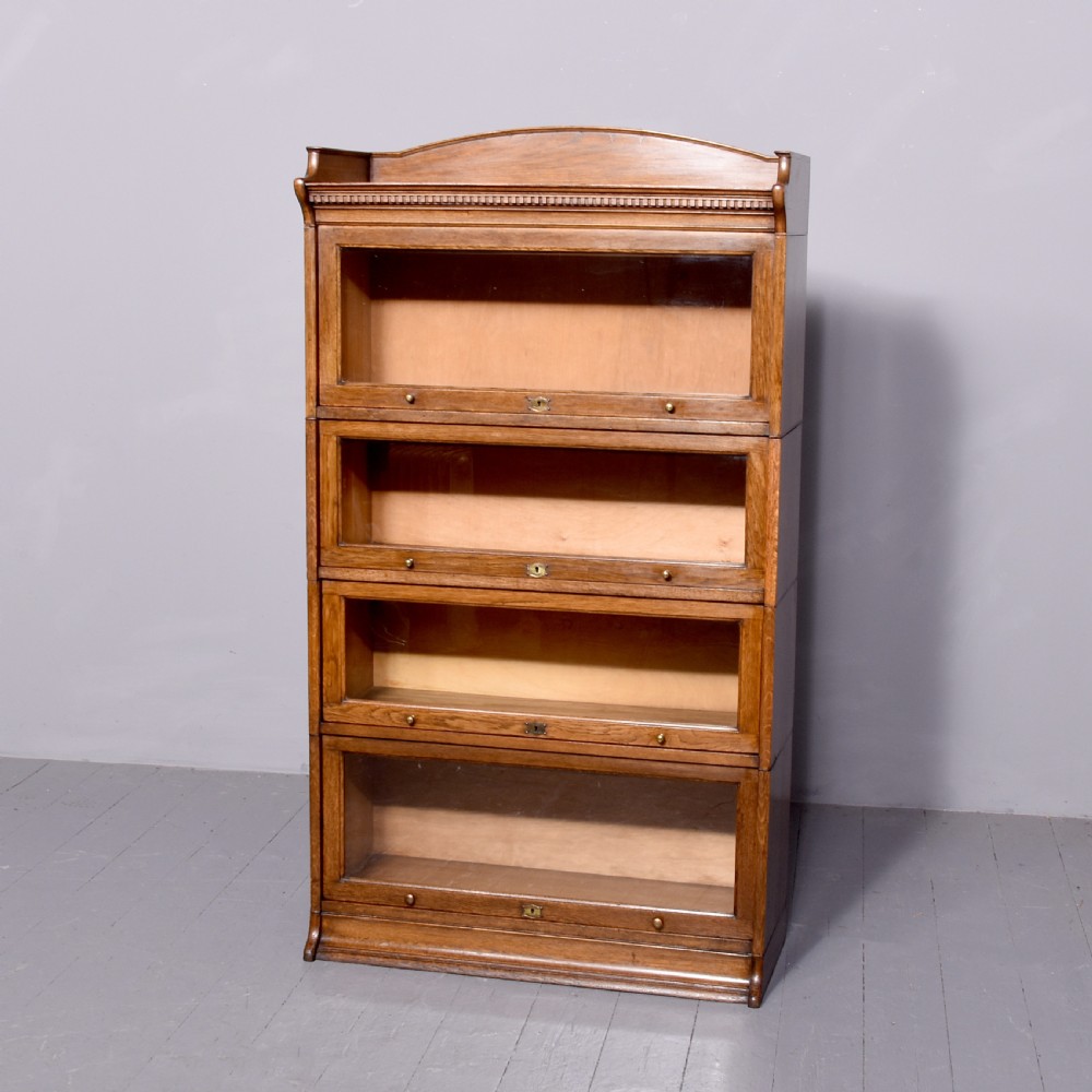 A four-tier Edwardian oak sectional bookcase featuring glazed up-and-over doors, brass handles, and an arched gallery top rail.