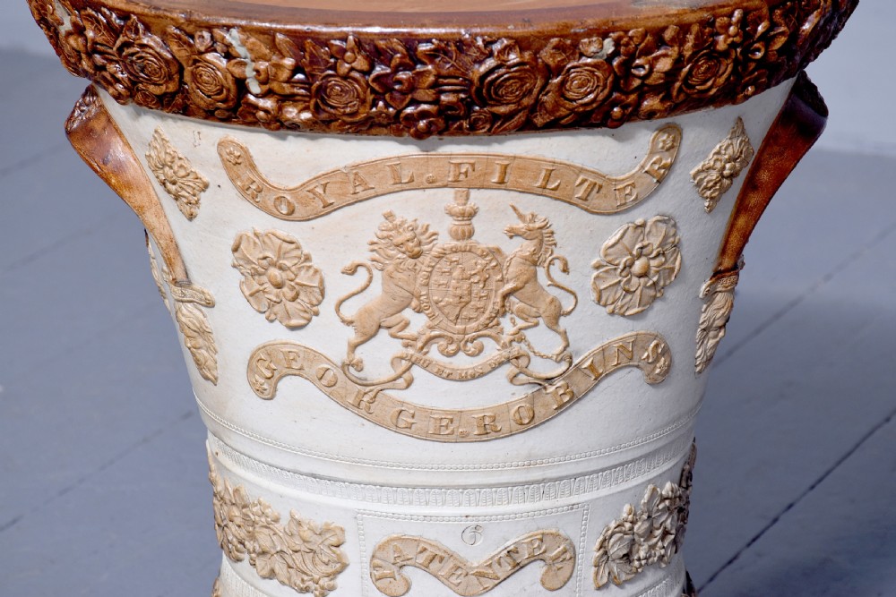 A Victorian glazed stoneware water filter urn decorated with relief crest motifs, floral borders, side handles, and a fitted wooden lid. View V