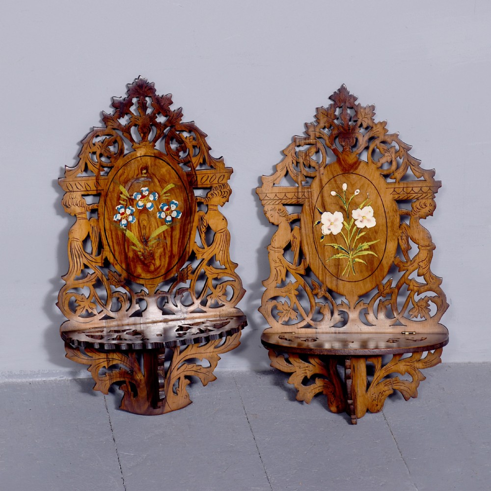 A pair of carved brown wooden wall brackets featuring pierced fretwork, foliage motifs, and oval central panels with painted floral designs.