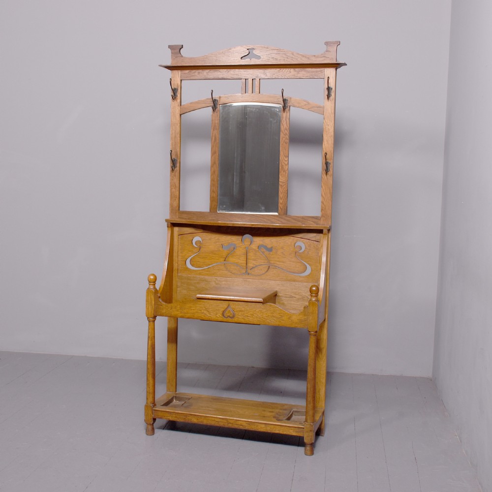 An antique oak Art Nouveau hall stand featuring a central mirror, brass coat hooks, a small shelf compartment, and a lower umbrella stand base.