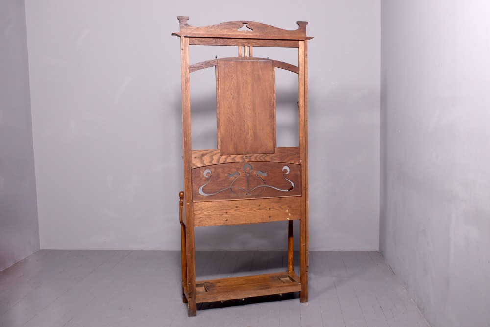 An antique oak Art Nouveau hall stand featuring a central mirror, brass coat hooks, a small shelf compartment, and a lower umbrella stand base. View II