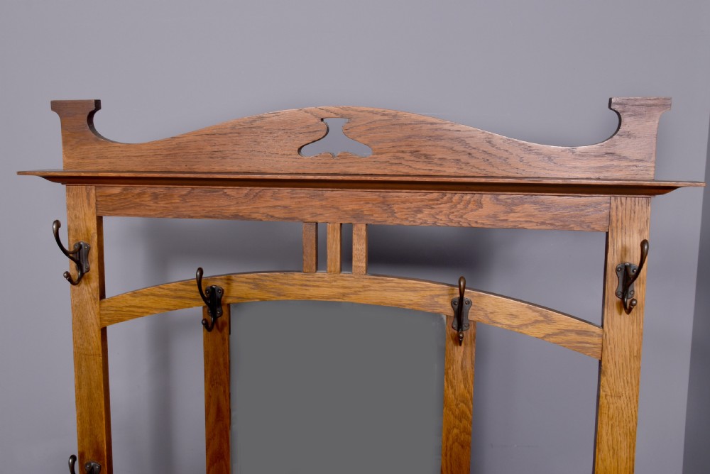 An antique oak Art Nouveau hall stand featuring a central mirror, brass coat hooks, a small shelf compartment, and a lower umbrella stand base. View III