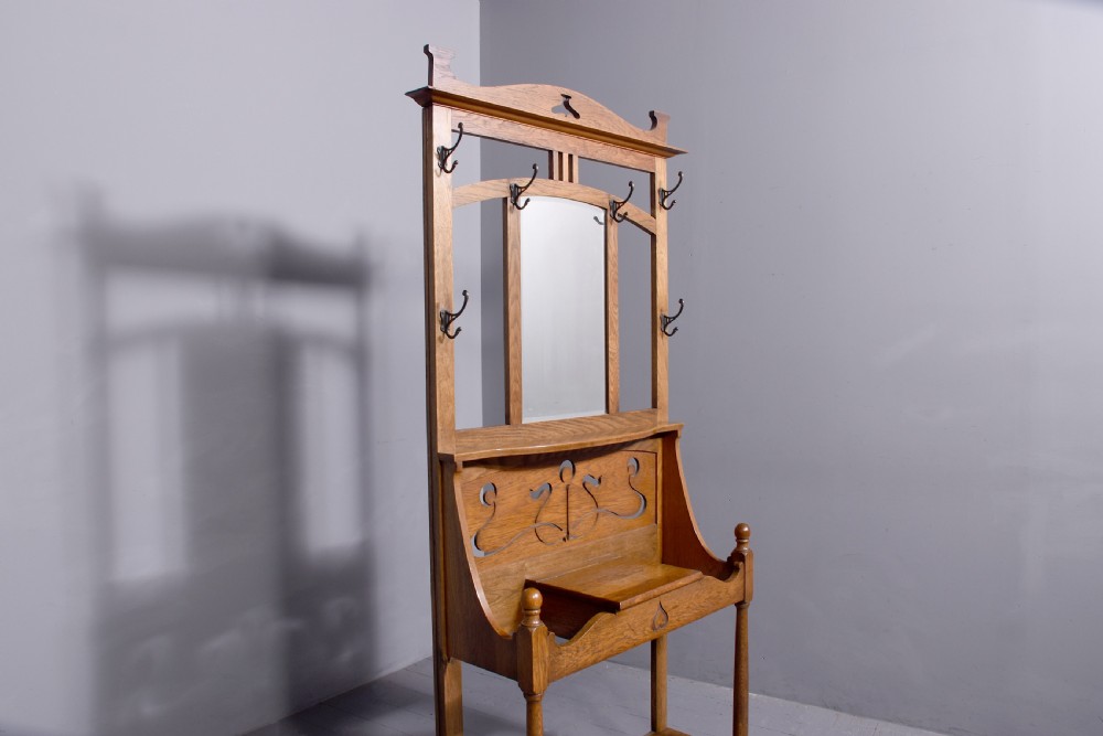 An antique oak Art Nouveau hall stand featuring a central mirror, brass coat hooks, a small shelf compartment, and a lower umbrella stand base. View IV