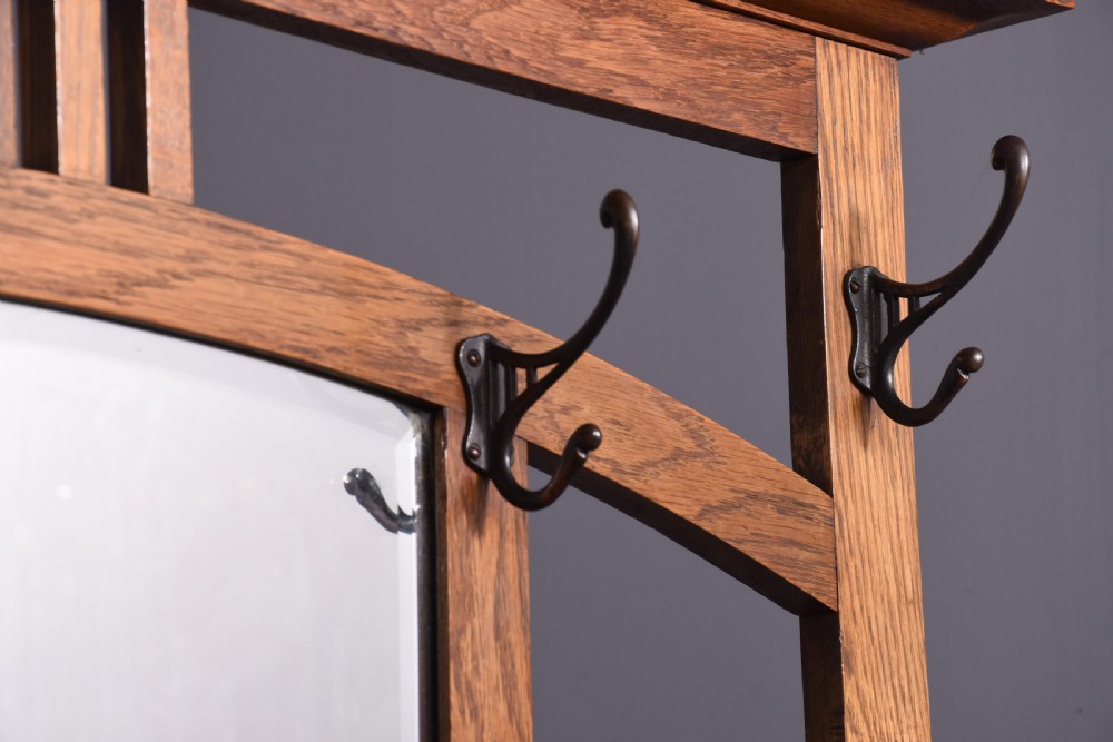 An antique oak Art Nouveau hall stand featuring a central mirror, brass coat hooks, a small shelf compartment, and a lower umbrella stand base. View VIII