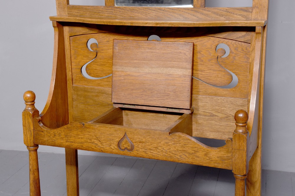 An antique oak Art Nouveau hall stand featuring a central mirror, brass coat hooks, a small shelf compartment, and a lower umbrella stand base. View IX
