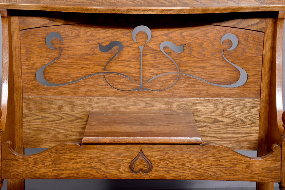 An antique oak Art Nouveau hall stand featuring a central mirror, brass coat hooks, a small shelf compartment, and a lower umbrella stand base. View X