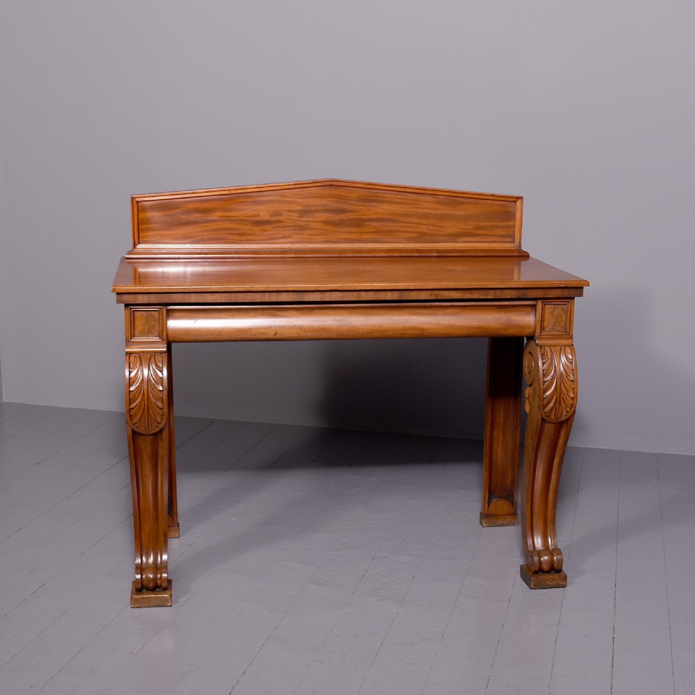 A William IV mahogany console table featuring a pedimented upstand, flame mahogany frieze, and acanthus leaf carved scroll front legs resting on plinth bases.