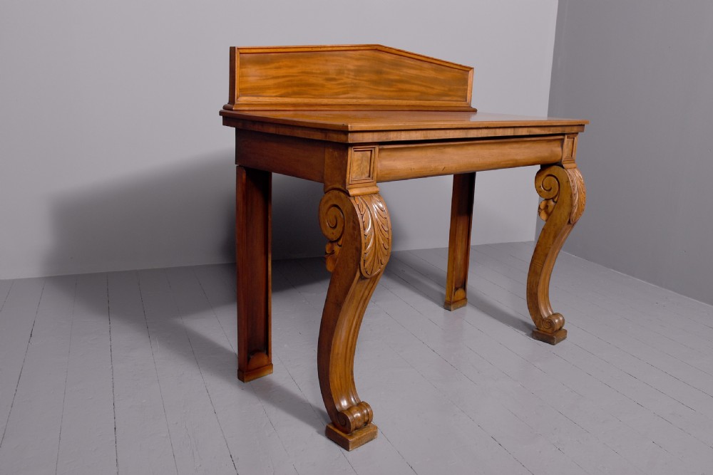 A William IV mahogany console table featuring a pedimented upstand, flame mahogany frieze, and acanthus leaf carved scroll front legs resting on plinth bases. View IV