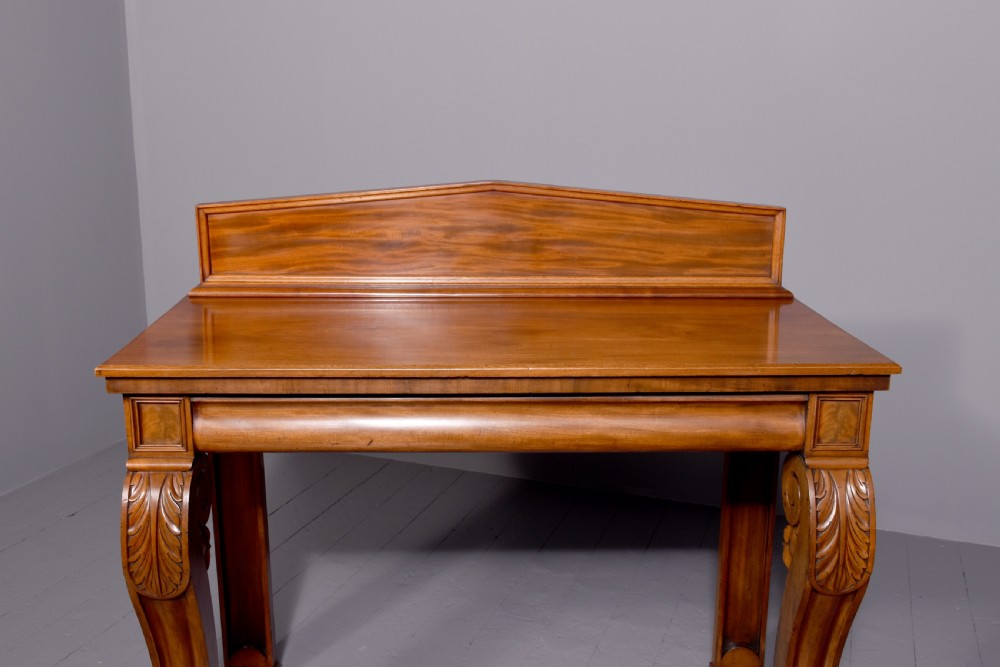 A William IV mahogany console table featuring a pedimented upstand, flame mahogany frieze, and acanthus leaf carved scroll front legs resting on plinth bases. View V