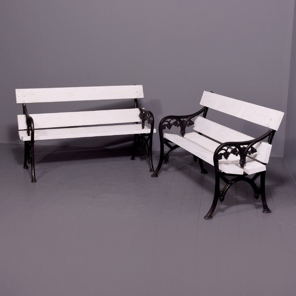 A pair of Victorian garden benches featuring dark cast iron side frames with ornate foliage scrollwork and white painted timber slatted seats and backs.