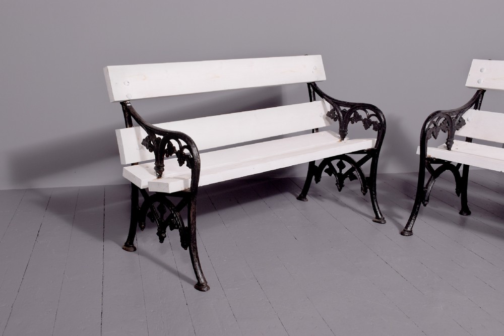 A pair of Victorian garden benches featuring dark cast iron side frames with ornate foliage scrollwork and white painted timber slatted seats and backs. View IV