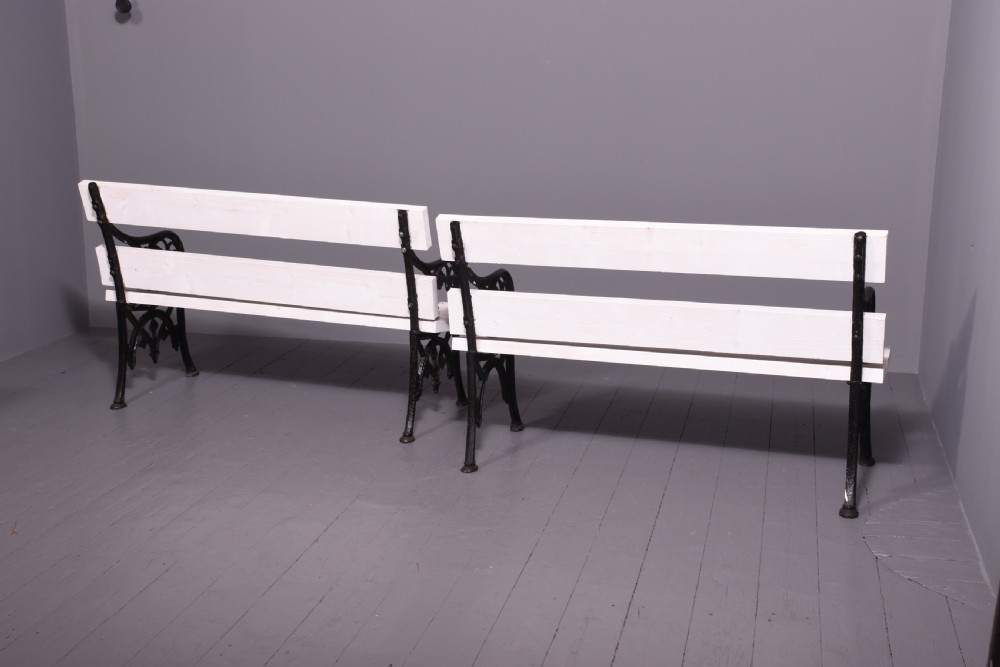 A pair of Victorian garden benches featuring dark cast iron side frames with ornate foliage scrollwork and white painted timber slatted seats and backs. View V