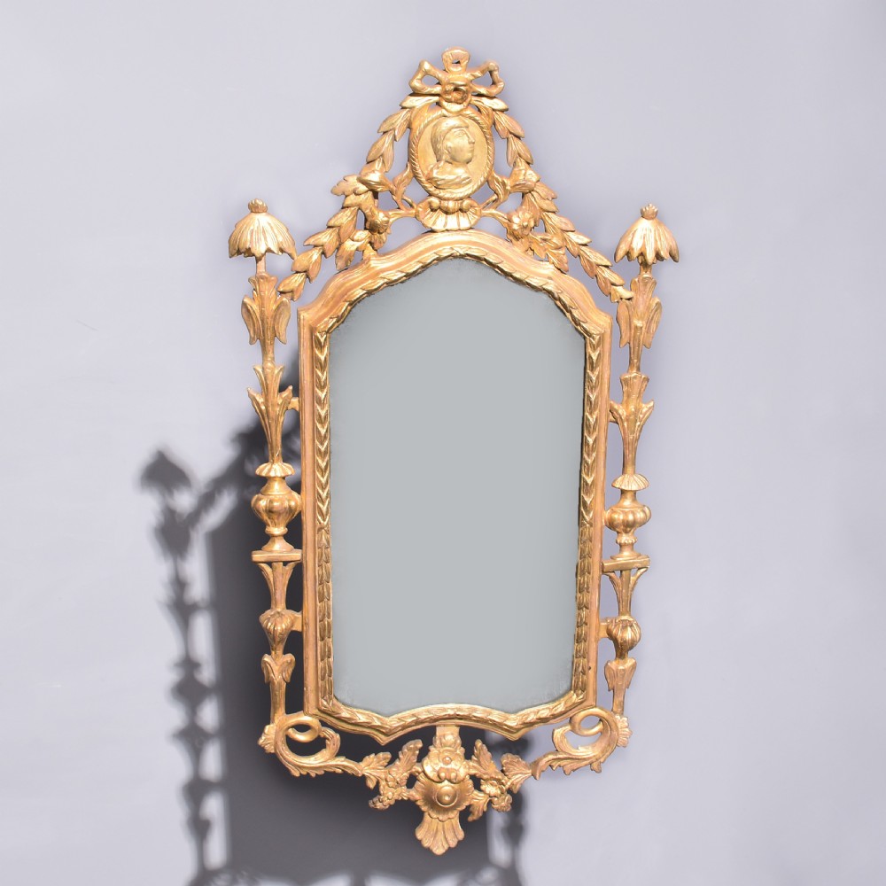 A late 18th-century Neoclassical giltwood wall mirror with a carved ribbon crest, profile medallion, and side foliate columns.