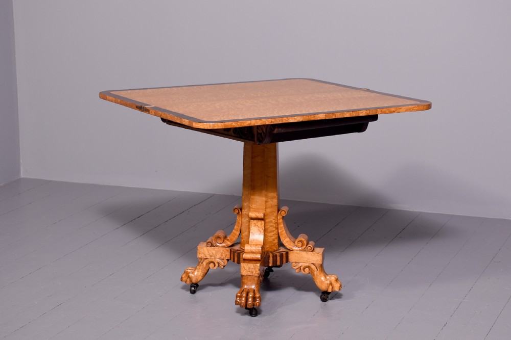 A George IV birdseye maple fold-over tea table featuring rosewood crossbanding, a central pedestal, and carved lion paw feet on brass castors.