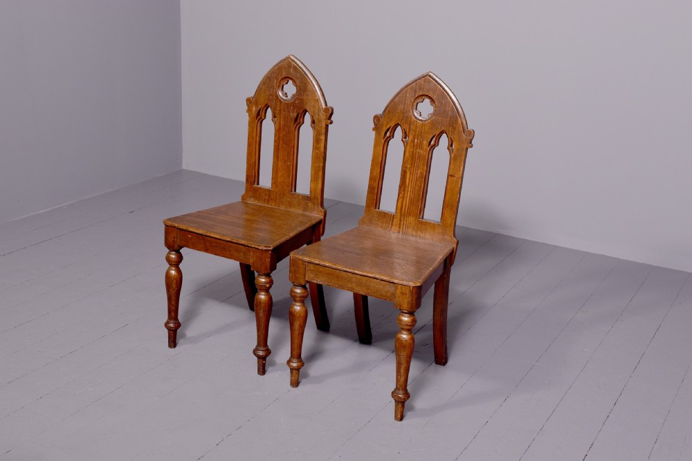 A pair of 19th-century pitch pine Gothic Revival hall chairs with pointed arch backrests, pierced trefoil motifs, turned front legs, and solid timber seats.