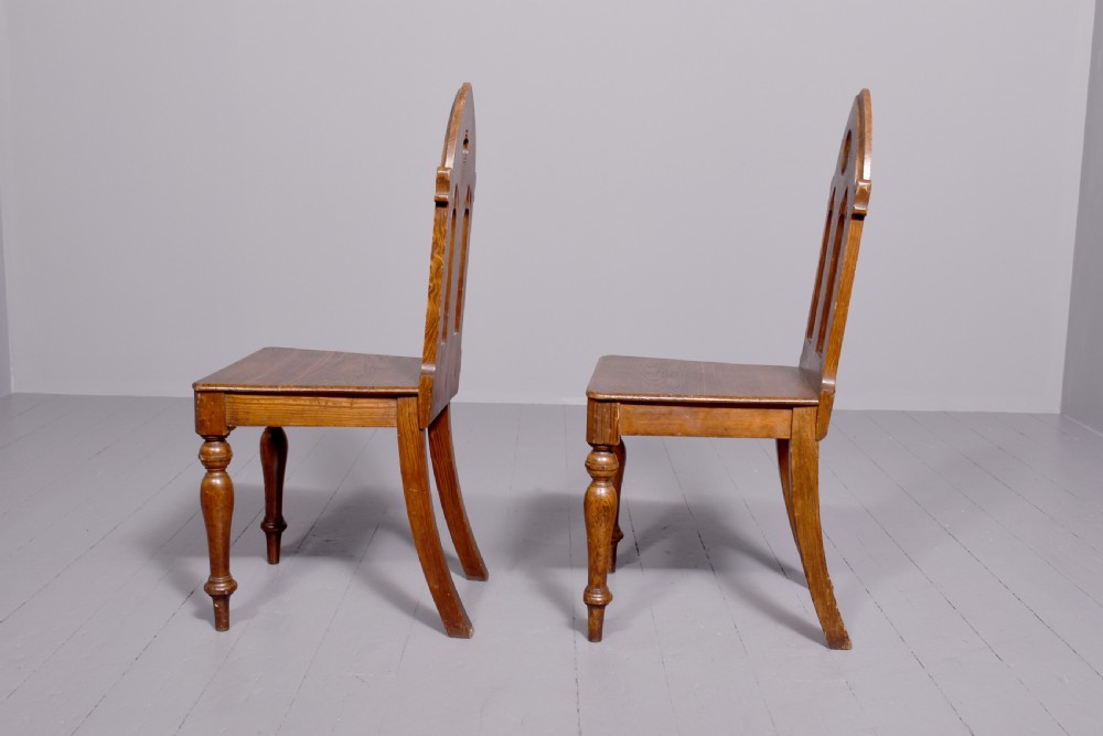 A pair of 19th-century pitch pine Gothic Revival hall chairs with pointed arch backrests, pierced trefoil motifs, turned front legs, and solid timber seats. View VI