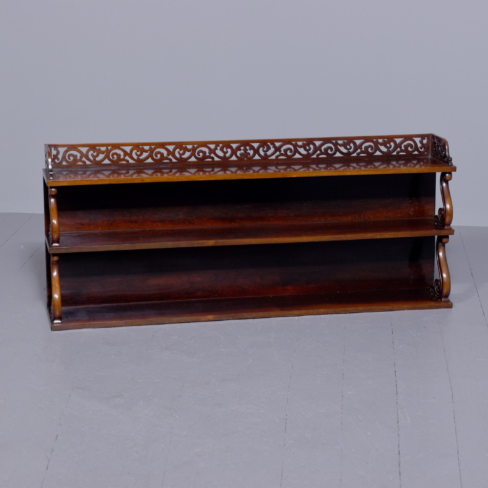 A three-tier William IV dark rosewood wall shelf featuring open shelving, curved S-scroll side supports, and a pierced fretwork top gallery rail.