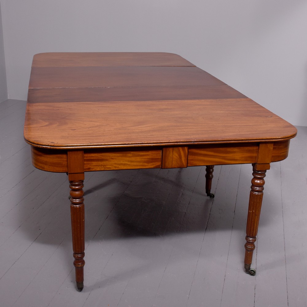 An extendable George IV mahogany dining table featuring a rectangular top with rounded corners, resting on four reeded turned legs with brass castors.