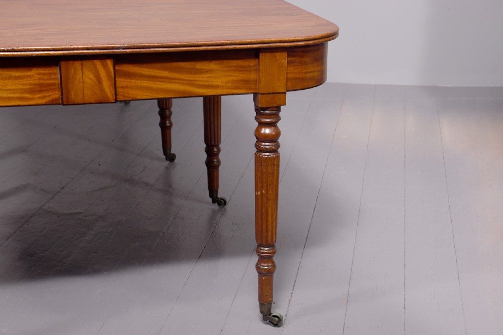 An extendable George IV mahogany dining table featuring a rectangular top with rounded corners, resting on four reeded turned legs with brass castors. View II
