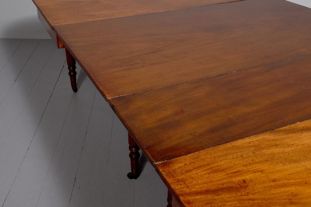 An extendable George IV mahogany dining table featuring a rectangular top with rounded corners, resting on four reeded turned legs with brass castors. View IV