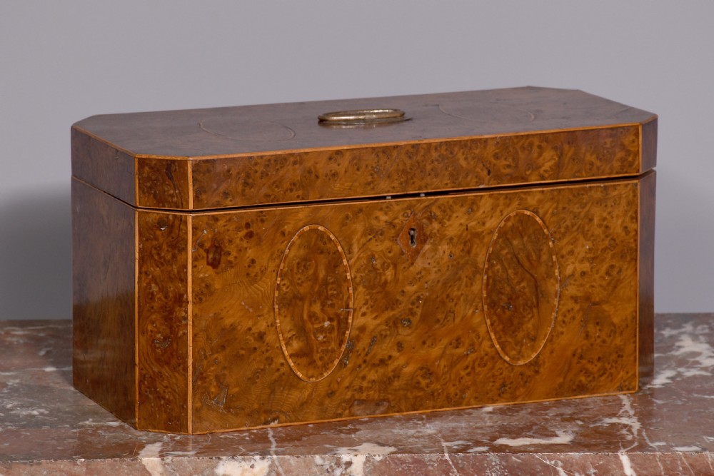 An 18th-century George III wooden tea caddy with a burr veneer finish, oval stringing inlays, canted corners, and a top brass handle.