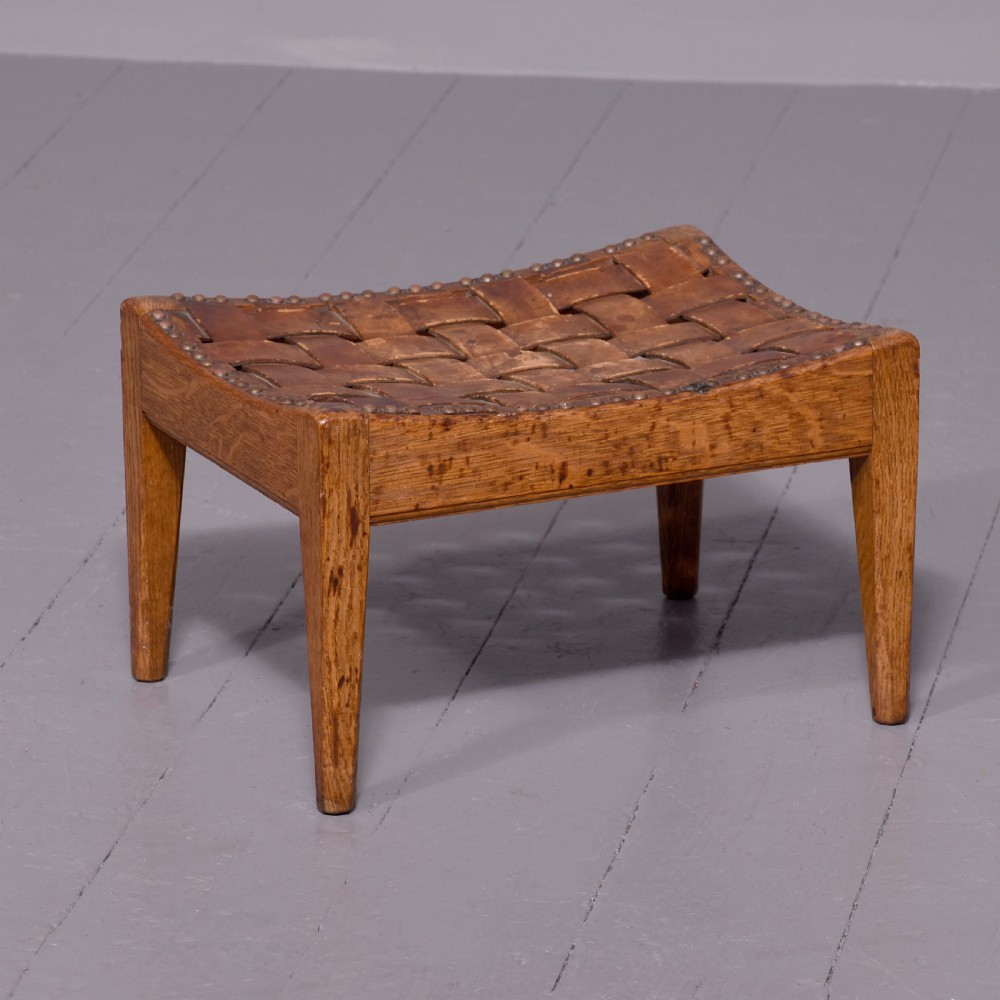 An Arts and Crafts oak footstool featuring a concave seat crafted from interwoven brown leather straps secured with metal studs, supported by four tapered square legs.