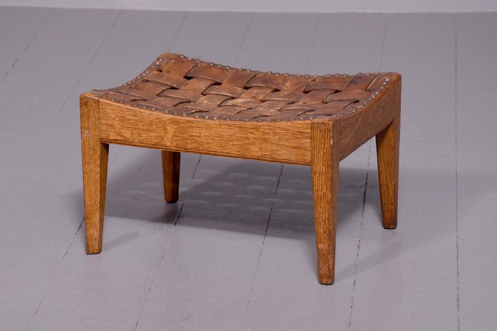 An Arts and Crafts oak footstool featuring a concave seat crafted from interwoven brown leather straps secured with metal studs, supported by four tapered square legs. View III