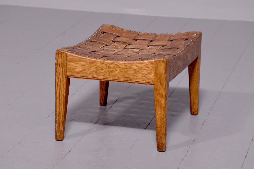 An Arts and Crafts oak footstool featuring a concave seat crafted from interwoven brown leather straps secured with metal studs, supported by four tapered square legs. View IV