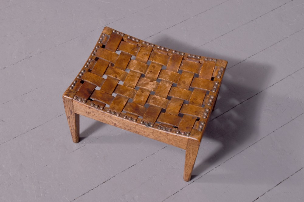 An Arts and Crafts oak footstool featuring a concave seat crafted from interwoven brown leather straps secured with metal studs, supported by four tapered square legs. View V