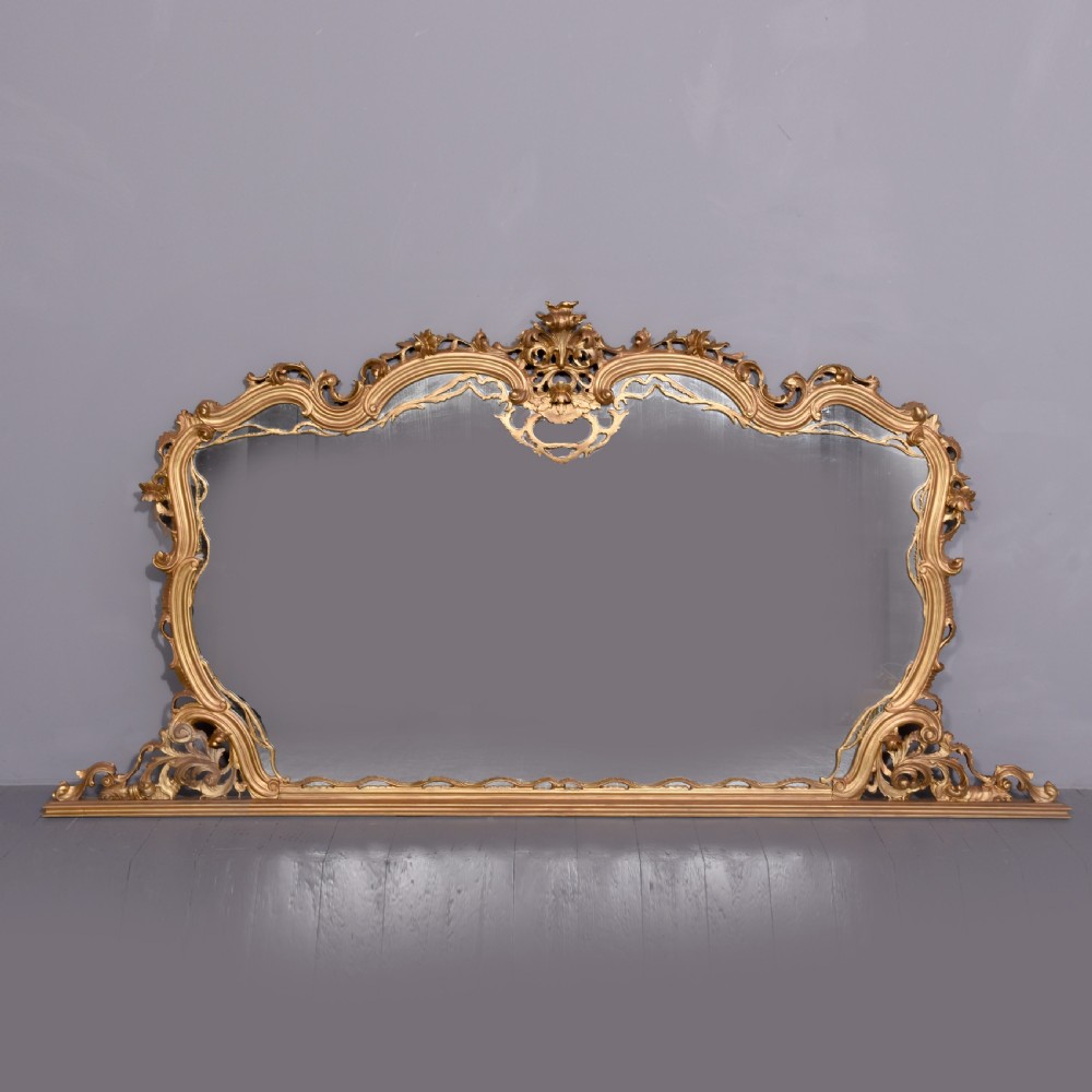 A wide carved giltwood overmantel mirror featuring ornate Rococo scrollwork, acanthus leaf motifs, and an arched cresting.