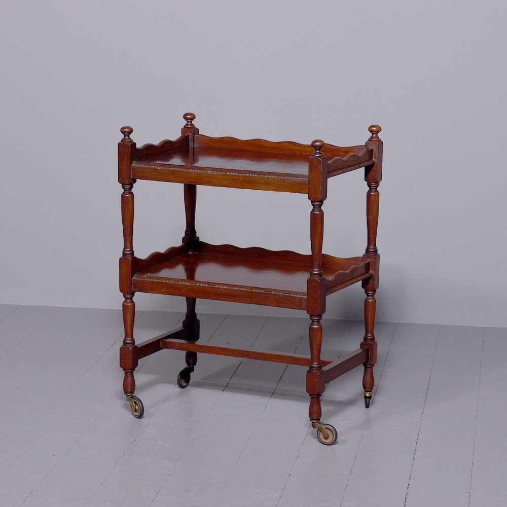 A two-tier mahogany wooden trolley featuring turned upright posts, brass castors, decorative inlay, and scalloped gallery borders.