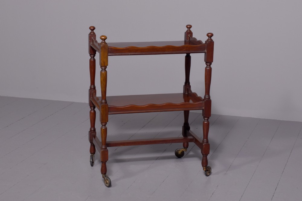 A two-tier mahogany wooden trolley featuring turned upright posts, brass castors, decorative inlay, and scalloped gallery borders. View IV