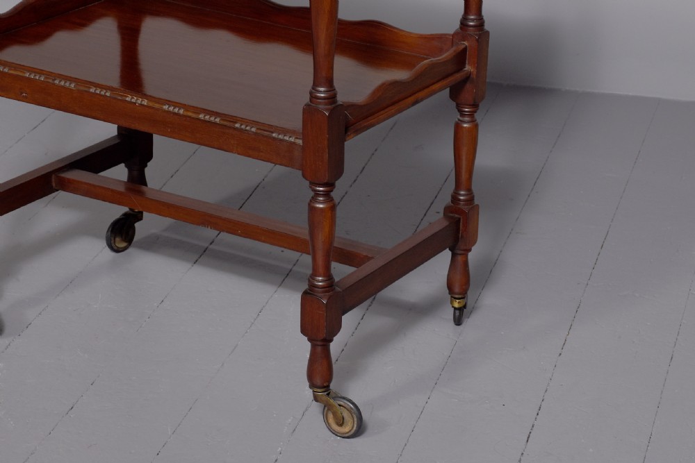 A two-tier mahogany wooden trolley featuring turned upright posts, brass castors, decorative inlay, and scalloped gallery borders. View V
