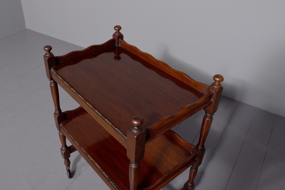 A two-tier mahogany wooden trolley featuring turned upright posts, brass castors, decorative inlay, and scalloped gallery borders. View VI