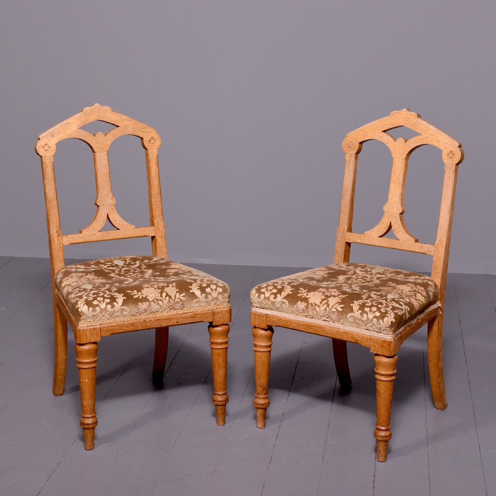 A pair of light oak side chairs featuring Gothic arched cut-outs on the backrests, turned front legs, and patterned gold velvet upholstery.