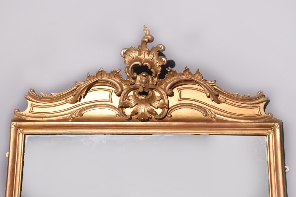 A large Victorian giltwood overmantle mirror featuring a curved top frame surmounted by a carved rococo cresting motif. View II