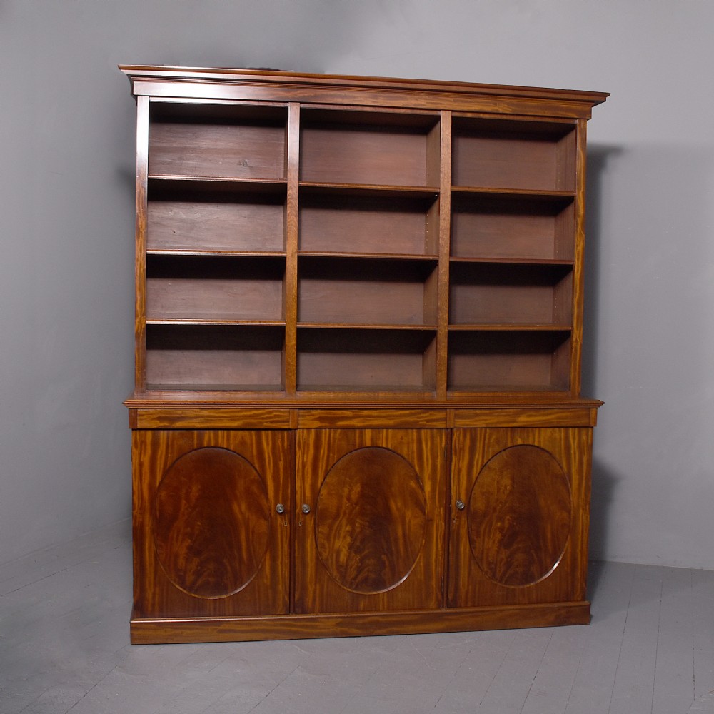 A large mid-Victorian mahogany bookcase featuring a three-bay open upper section with adjustable shelves set above a three-door cupboard base with oval veneered panels.