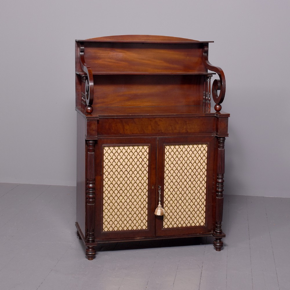 A dark mahogany Regency chiffonier featuring an upper open shelf supported by scrolled sides and two lower cupboard doors with diamond brass grilles.