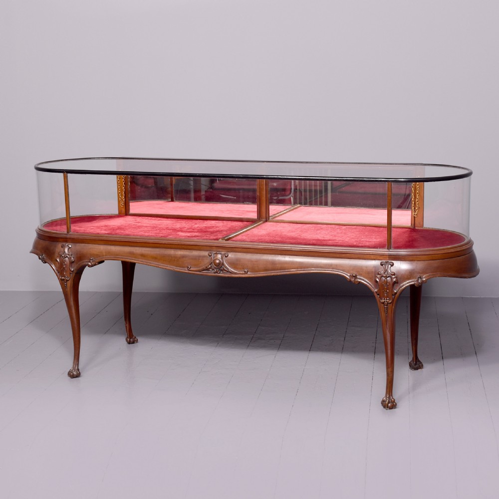 A large oval mahogany display cabinet featuring curved glass sides, red velvet lining, carved cabriole legs, and claw and ball feet.