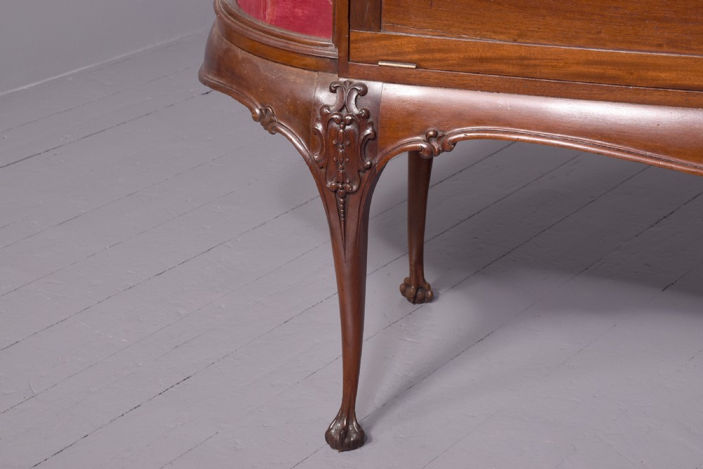 A large oval mahogany display cabinet featuring curved glass sides, red velvet lining, carved cabriole legs, and claw and ball feet. View XIII