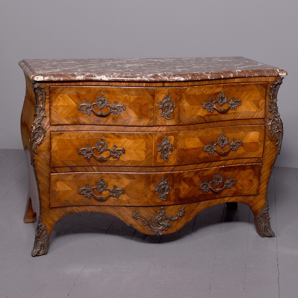 A Louis XV style bombee commode featuring a variegated marble top, geometric timber parquetry, three drawers, and ornate bronze handles.