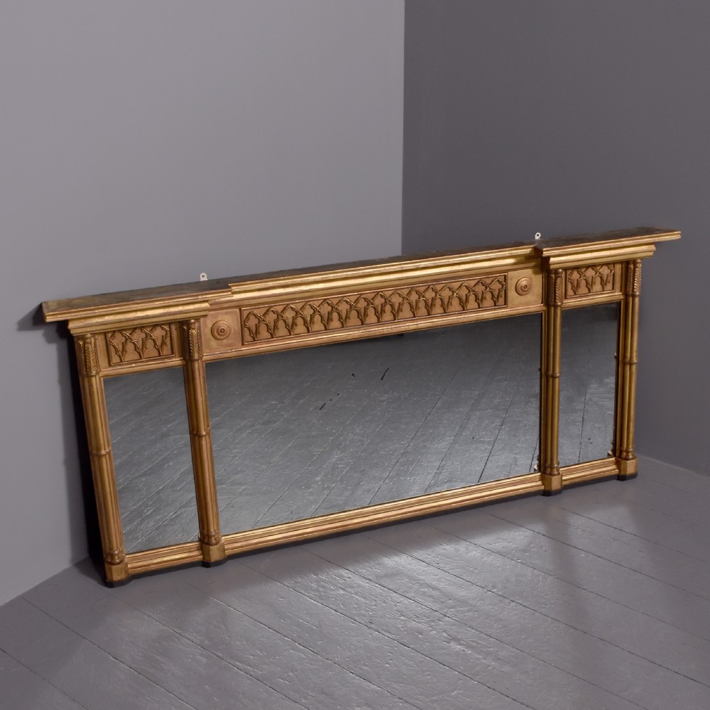 A wide Regency giltwood tryptic overmantel mirror featuring three mirror plates, carved blind tracery frieze, and reeded upright columns.
